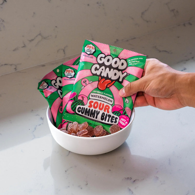 Hand holding a bag of Good Candy Watermelon low sugar sour gummies above a white bowl of gummy candies on a marble countertop