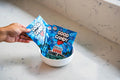 Hand holding two bags of Good Candy Blue Razz low sugar sour gummies above a white bowl on a marble countertop