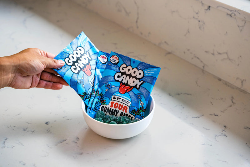 Hand holding two bags of Good Candy Blue Razz low sugar sour gummies above a white bowl on a marble countertop